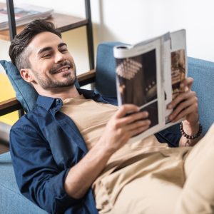 Happy young man with beard is lying on sofa and reading magazine. He is holding journal in both hands and smiling